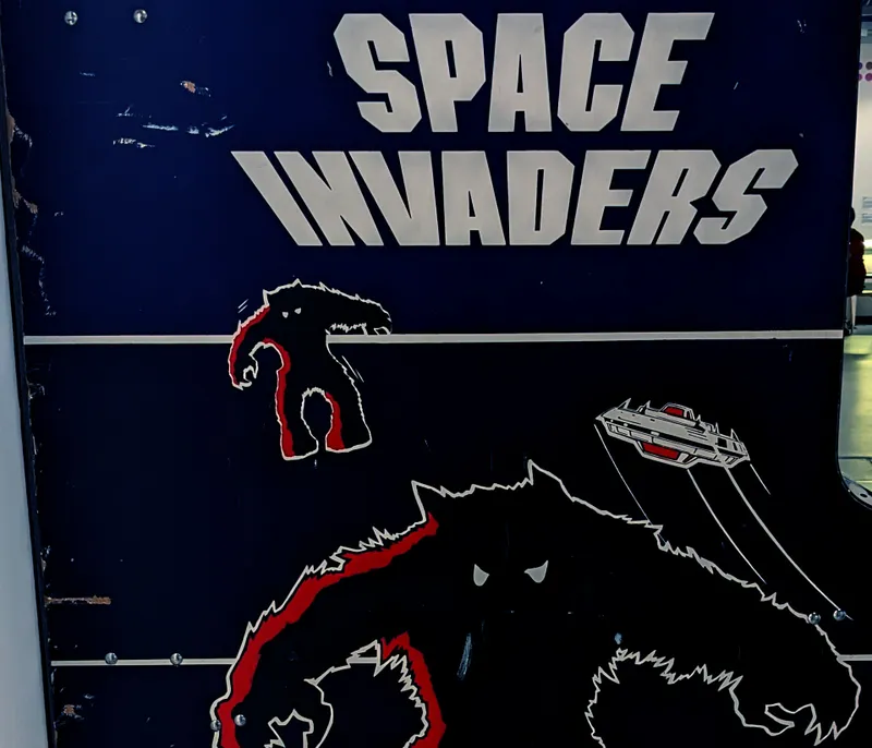 A close-up of an arcade cabinet featuring the classic game "Space Invaders." The cabinet is predominantly dark blue, with bold white lettering spelling out "SPACE INVADERS" at the top. Below, stylized graphics depict a menacing alien creature in black and red, along with a spaceship firing projectiles, capturing the retro aesthetic of early video gaming. The overall design evokes nostalgia for the arcade era.