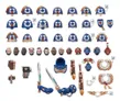 A collection of Ultramarine upgrade components is displayed, featuring various sculpted shoulder pads, helmets, and accessories in a blue and gold color scheme. The parts include multiple shoulder pads designed for different unit types, as well as heads, swords, and decorative elements like laurels and purity seals. Additionally, a transfer sheet with numerous decals for customization is included, showcasing campaign badges and Ultramarines Chapter icons.