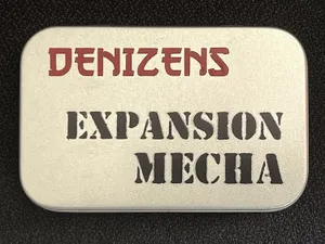 A small, silver tin features the text "DENIZENS" in bold red letters at the top and "EXPANSION MECHA" in large, black, stencil-style font below. The tin's surface has a smooth, metallic finish, and it is set against a dark background, emphasizing its compact and portable design. This packaging represents the new expansion for the Space Denizens wargame, which includes mechs and terrain features.