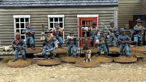 A group of ten detailed 28mm scale miniatures representing dismounted US Cavalry and Buffalo Soldiers stands in front of a wooden building with a red door. The miniatures are posed with various weapons, including rifles and a bugle, showcasing a mix of uniforms and accessories, such as scarves and hats. A small beagle and a cactus are also present among the figures, all mounted on 25mm round bases with a sandy terrain effect.