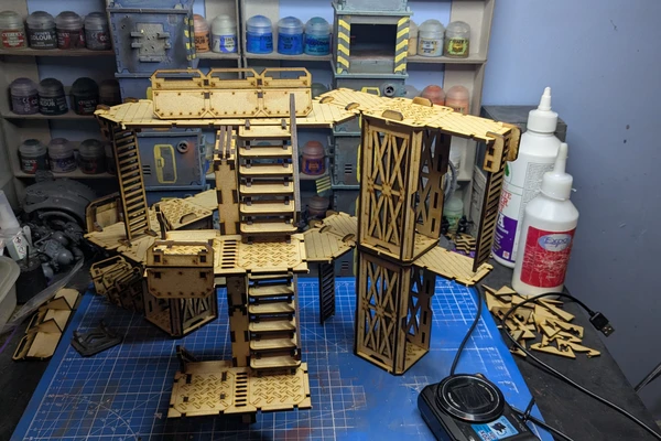 A detailed assembly of modular terrain pieces made from laser-cut wood, featuring multiple levels, staircases, and platforms. The structure includes various textures and patterns, with a mix of open and enclosed areas, showcasing a futuristic industrial design. In the background, paint bottles and tools are visible, indicating a workspace for painting and customizing the terrain.