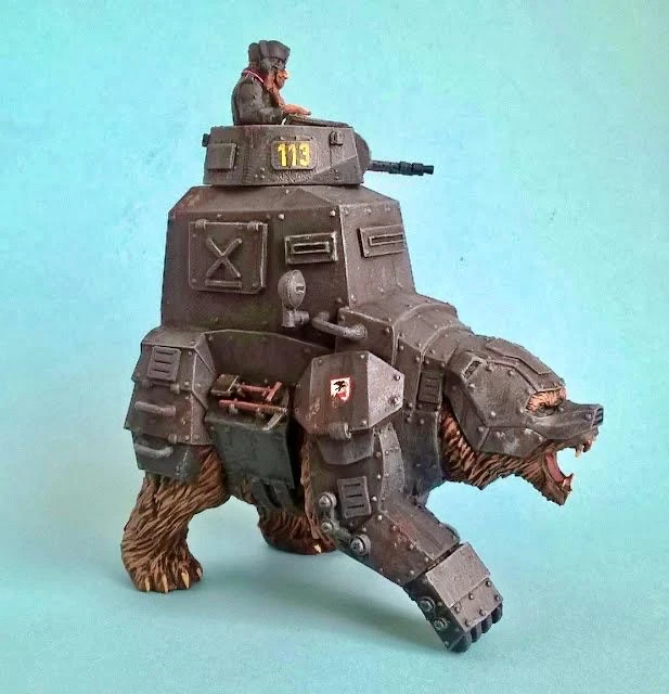 A detailed miniature depicts a mechanized bear, armored with various metal plates and featuring a turret on its back. A small figure, dressed in military attire, sits atop the turret, overseeing the bear's movement. The bear's fierce expression and the intricate design of the armor highlight the blend of fantasy and military themes characteristic of the Panzerfäuste game.