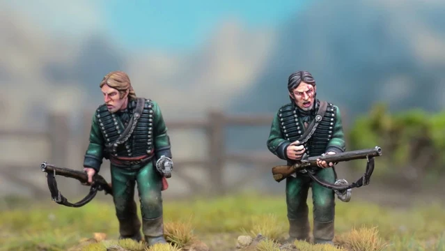 Two detailed miniatures of soldiers from the Sharpe’s Rifles series are displayed on textured bases, set against a blurred natural background. Both figures wear green uniforms with black crossbelts; one holds a rifle and has a serious expression, while the other appears to be in a more contemplative pose. The bases feature tufts of grass, enhancing the realistic terrain effect.
