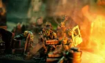 A group of colorful, exaggerated space orc miniatures is depicted amidst a chaotic urban battlefield, surrounded by piles of debris and scattered weapons. One orc, wearing goggles and a top hat, holds a large gun, while others are engaged in various antics, including wielding makeshift weapons. The scene is illuminated with a warm, fiery glow, enhancing the chaotic and humorous atmosphere of the skirmish game.