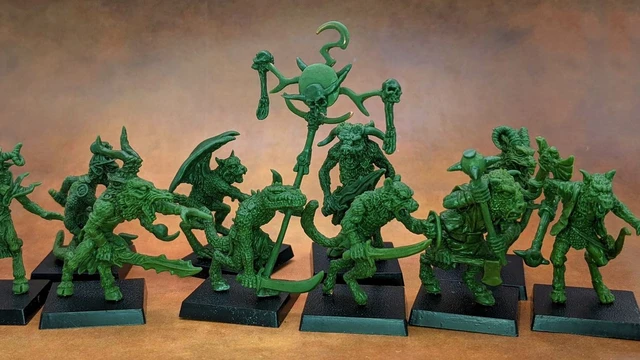 A collection of old school miniatures from Satyr Art Studio, featuring various fantastical creatures in a vibrant green color. The figures include beastmen with distinct features such as horns and claws, some wielding weapons, and a central figure holding a staff topped with a skull and a crescent. They are displayed on black bases against a warm, textured background.
