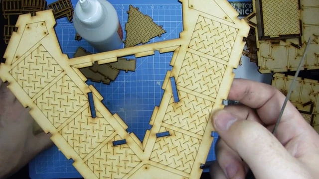 A close-up view of a partially assembled model from the Slum Shanties set by TT Combat, showcasing intricate details like diamond plate textures on the wooden pieces. In the foreground, a hand holds a section of the model, while various cut-out pieces are visible on a blue cutting mat in the background. A bottle of glue is also present, indicating the assembly process.