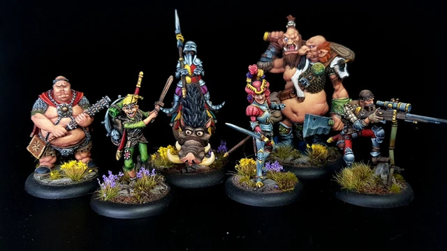A group of eight intricately painted Moonstone miniatures stands against a black background. The characters include a muscular figure with a weapon, a green-skinned goblin in colorful attire, a knight in armor, and various other fantastical beings, each displaying unique features and vibrant colors. They are positioned on circular bases adorned with tufts of grass and small flowers, showcasing the detailed artistry of the miniatures.