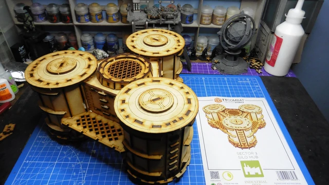 A detailed model of the Sector 3 Silo Hub from TT Combat is prominently displayed on a blue cutting mat. The structure features multiple cylindrical silos with intricate wooden textures and circular tops, showcasing a combination of open latticework and solid panels. In the background, various paint pots and a few other miniature models are visible, along with the instruction sheet for the Silo Hub, which includes an illustration of the assembled model.