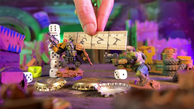 A vibrant tabletop scene features two miniatures engaged in a skirmish, surrounded by scattered dice and bottle caps. One miniature, a heavily armed space orc, stands confidently while a second, more humanoid figure aims a weapon. A hand holds a ruler above the orc, suggesting a measurement, with a colorful, chaotic backdrop of painted terrain and additional game elements enhancing the playful atmosphere.