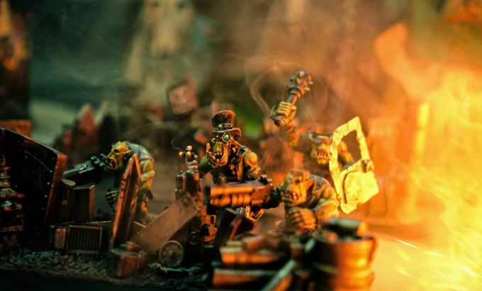 A group of colorful, exaggerated space orc miniatures is depicted amidst a chaotic urban battlefield, surrounded by piles of debris and scattered weapons. One orc, wearing goggles and a top hat, holds a large gun, while others are engaged in various antics, including wielding makeshift weapons. The scene is illuminated with a warm, fiery glow, enhancing the chaotic and humorous atmosphere of the skirmish game.