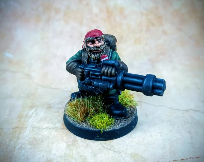 A detailed miniature of a space dwarf crouches on a round base adorned with tufts of green grass. The dwarf has a bushy beard, wears a red cap, and is dressed in a green jacket with a backpack, holding a large, futuristic weapon. The background features a textured, neutral surface that enhances the focus on the miniature.