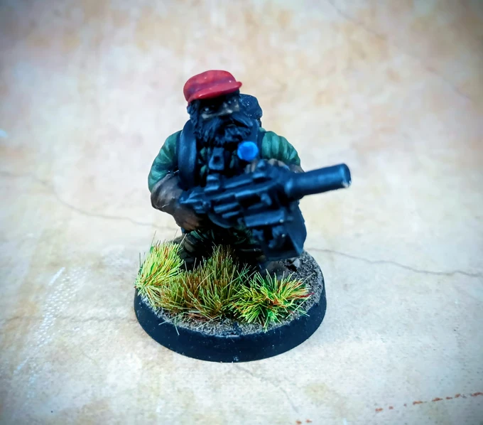 A miniature figure of a space dwarf jungle operative is depicted, crouching and holding a futuristic weapon. The figure wears a red cap and a green jacket, with a dark beard and a determined expression. The base is adorned with tufts of grass, set against a textured, neutral background.