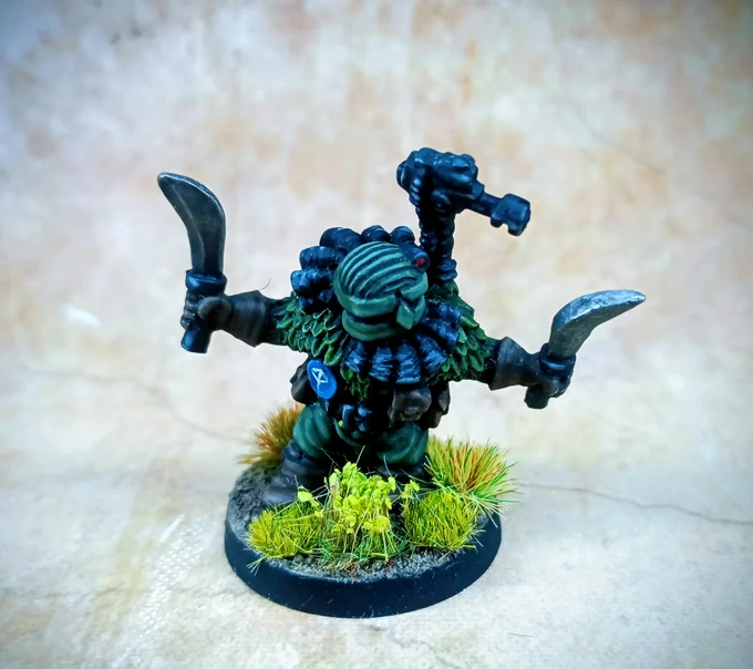 A jungle operative miniature stands on a textured base adorned with tufts of green grass. The figure is clad in dark green armor with a distinctive feathered collar, holding two curved blades in its hands. A small, mechanical device is mounted on its back, and a red eye is visible on its head, adding to its menacing appearance.