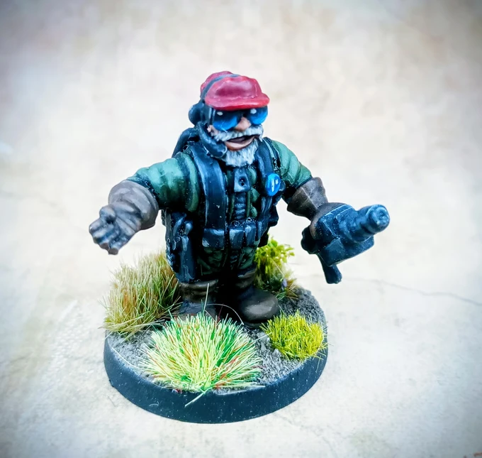 A miniature figure of a space dwarf stands on a textured base adorned with tufts of green grass. The dwarf has a bushy beard, wears a red cap, and sports blue goggles, along with a green and black tactical outfit. He is holding a futuristic weapon in one hand and extending the other as if gesturing or calling out.