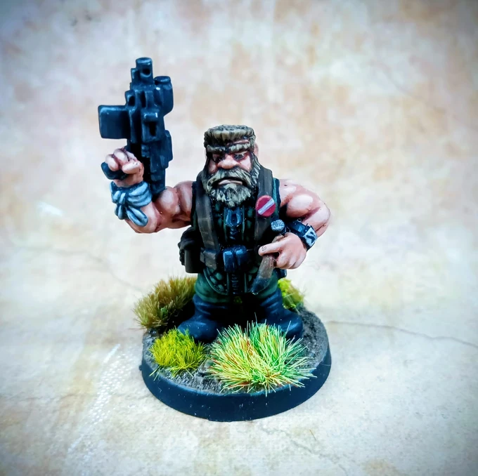 A muscular, bearded miniature figure stands confidently, holding a futuristic firearm in one hand and gesturing with the other. He wears a tactical vest and has a rugged appearance, complete with a bandana and various accessories attached to his outfit. The base features tufts of green grass, enhancing the character's jungle operative theme against a textured, neutral background.