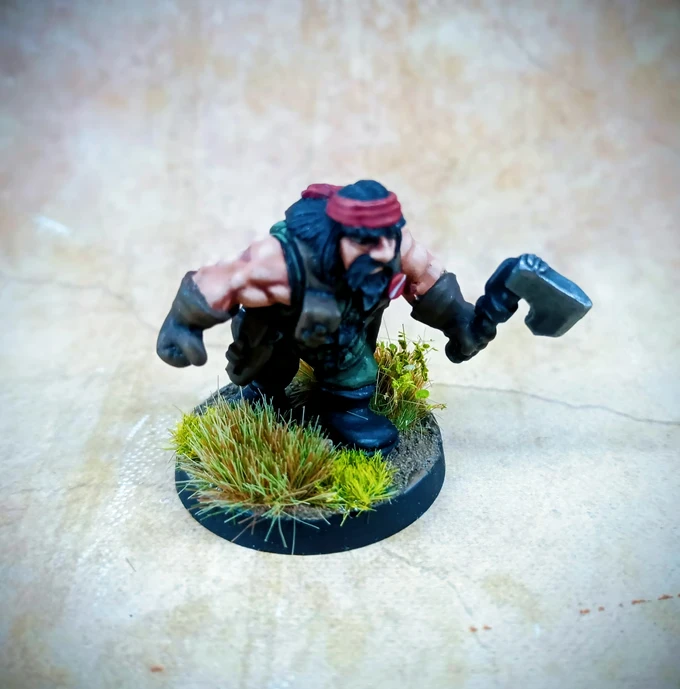 A muscular, bearded dwarf miniature is depicted in a dynamic pose, wielding a large axe in one hand while crouching slightly. He wears a green vest and dark pants, with a red bandana around his head and fingerless gloves. The base is adorned with tufts of grass, enhancing the jungle theme, against a textured, neutral background.