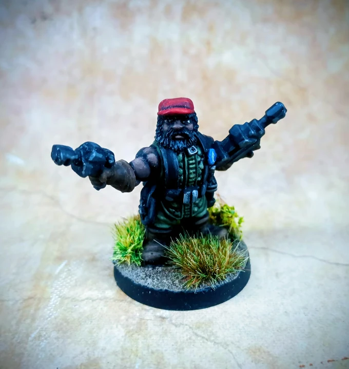 A miniature figure of a space dwarf stands confidently, holding a weapon in each hand. The dwarf has a thick beard and wears a red cap, along with a green tactical outfit adorned with various pouches. The base is detailed with tufts of grass and a textured surface, enhancing the overall appearance of the figure.