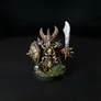 A miniature figure of a Chaos Dwarf warrior with a horned helmet, holding a large round shield with a spiked boss on the left arm and a sword in the right hand. The figure has a long braided beard, detailed armor, and is standing on a small circular base with patches of grass. The background is black.