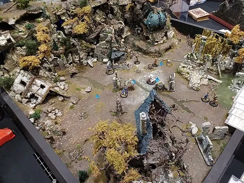 A detailed tabletop gaming diorama showcases a rugged, rocky terrain with scattered ruins and overgrown vegetation. The landscape features a mix of rocky outcrops, broken structures, and patches of yellow foliage, creating a post-apocalyptic atmosphere. Small miniatures are positioned throughout the scene, suggesting an ongoing game or battle among the ruins.