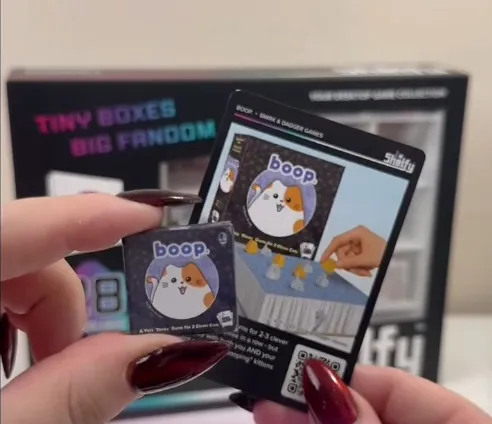 A hand with red nail polish holds two small game boxes labeled "boop." The box in focus has a cartoon cat with orange and white fur and a purple background. The other box shows an image of a hand placing a yellow token on a game board. In the background, a display shelf holds multiple similar small game boxes, with a sign above reading "TINY BOXES BIG FANDOM" and another sign reading "SHELFY."