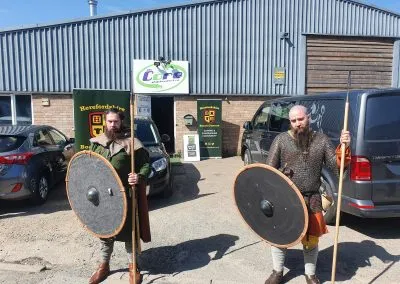 Two individuals dressed in historical warrior attire stand outside a building, each holding a round shield and a spear. They wear chainmail and tunics, with one having a long beard and the other a shorter beard. In the background, a sign for "Herefordshire Mind" is visible, along with a few parked cars and a large van.