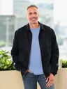 A man in a black jacket over a blue shirt, smiling at the camera. Office setting with modern glass windows and indoor plants in the background.