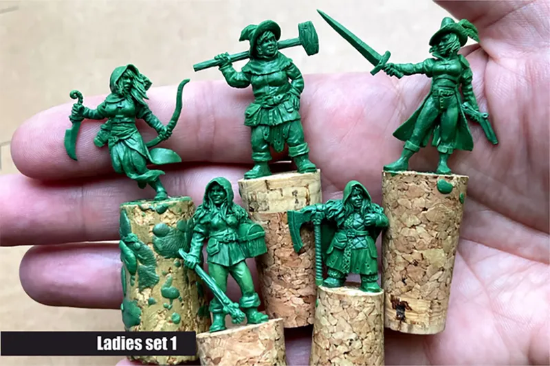 A hand holding several green resin miniatures from the "Ladies Only" collection by Scibor Monstrous Miniatures, displayed on cork bases. The miniatures depict various fantasy female characters, including a warrior with a sword, a rogue with a bow, and a dwarf wielding an axe, showcasing diverse poses and details. The text "Ladies set 1" is visible at the bottom of the image.