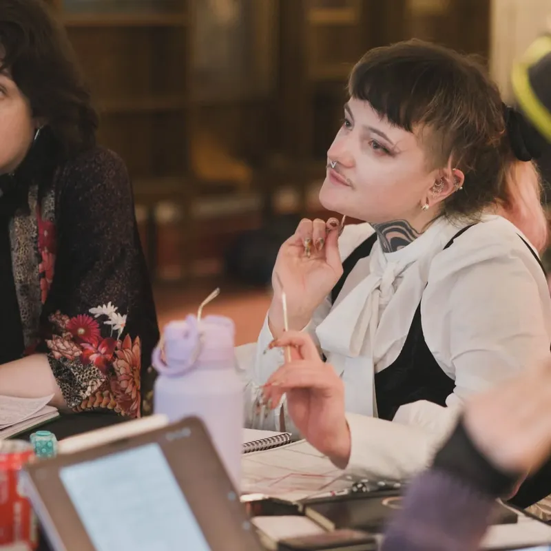 A group of individuals engaged in a tabletop gaming session, with one person in the foreground wearing a white blouse and sporting distinctive tattoos and facial piercings. They appear to be deep in thought, holding a pencil and looking attentively at the table. The setting features a mix of gaming materials, including dice, notebooks, and a water bottle, with a backdrop of a gothic-style library interior.
