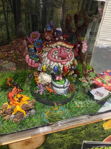 A whimsical, colorful miniature scene features a jovial gnome with a large, mushroom-shaped hat, surrounded by vibrant flowers and small creatures. The gnome holds a bowl and stands near a cauldron, with a campfire made of logs and flames in the foreground. The base is adorned with lush green grass and various decorative elements, creating a lively and enchanting atmosphere.