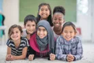 A group of six children is smiling and posing together on a light-colored carpet. They are arranged in a semi-circle, with the girl in the front wearing a pink sweater and a grey hijab, while the others are dressed in various colorful outfits. The background features a softly blurred interior space, suggesting a warm and welcoming environment.