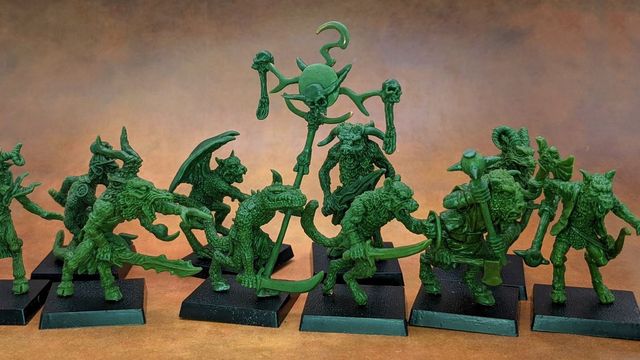 A collection of old school miniatures from Satyr Art Studio, featuring various fantastical creatures in a vibrant green color. The figures include beastmen with distinct features such as horns and claws, some wielding weapons, and a central figure holding a staff topped with a skull and a crescent. They are displayed on black bases against a warm, textured background.