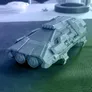 A detailed 3D-printed model of a futuristic vehicle, featuring a sleek, angular design with prominent headlights and various mechanical components. The model is displayed on a textured surface, with a blurred background that includes a circular object and some indistinct items. The overall color scheme is a monochromatic gray, highlighting the intricate details of the vehicle's structure.