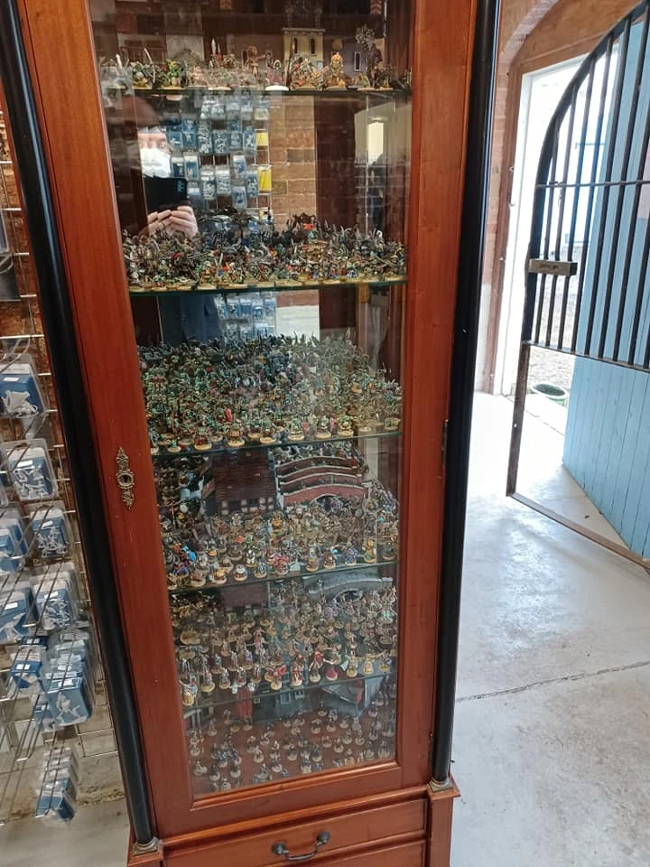 A wooden display cabinet filled with a vast array of painted miniatures, showcasing various fantasy figures and models. The cabinet has multiple shelves, each densely packed with detailed miniatures, including warriors, creatures, and vehicles, reflecting a rich history of tabletop gaming. In the background, a glimpse of the shop's interior and an open door can be seen, adding to the atmosphere of the Foundry's headquarters.