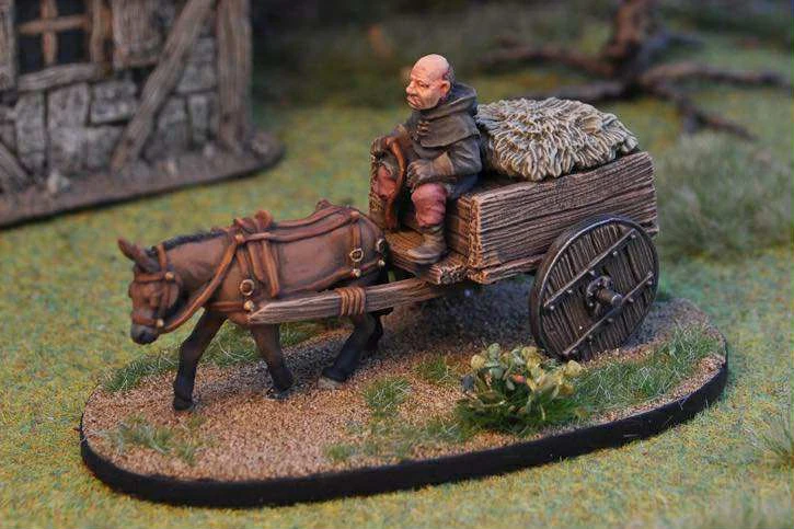 A miniature scene features a small cart being pulled by a brown horse, with a seated figure dressed in rustic clothing, holding the reins. The cart is laden with a bundle of hay or straw, and the base is detailed with grass and small plants. In the background, a stone building adds to the fantasy setting, enhancing the overall atmosphere of a quaint village scene.