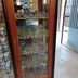 A wooden display cabinet filled with a vast array of painted miniatures, showcasing various fantasy figures and models. The cabinet has multiple shelves, each densely packed with detailed miniatures, including warriors, creatures, and vehicles, reflecting a rich history of tabletop gaming. In the background, a glimpse of the shop's interior and an open door can be seen, adding to the atmosphere of the Foundry's headquarters.