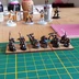 A collection of painted 10mm Orc miniatures is displayed on a green cutting mat, showcasing various poses and weapons. The Orcs are arranged on a wooden base, with some holding spears and shields, while others wield axes and a banner. In the background, partially visible models and tools suggest an active painting workspace, with bottles and a pink cutting mat adding to the scene's hobbyist atmosphere.