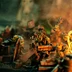 A group of colorful, exaggerated space orc miniatures is depicted amidst a chaotic urban battlefield, surrounded by piles of debris and scattered weapons. One orc, wearing goggles and a top hat, holds a large gun, while others are engaged in various antics, including wielding makeshift weapons. The scene is illuminated with a warm, fiery glow, enhancing the chaotic and humorous atmosphere of the skirmish game.