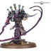 A detailed miniature of a character from the Emperor’s Children faction stands prominently, showcasing intricate design elements. The figure features a pale, skull-like face and is adorned with dark armor, highlighted by vibrant purple and green accents, including tubes on the back. In one hand, it wields a curved blade, while a long, whip-like appendage extends from the other side, all set on a textured base with scattered rocks and grass.