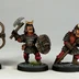 Three fantasy miniatures are displayed, each depicting a character in armor with distinct features. The first figure on the left holds a bow, while the middle figure wields an axe and a shield, showcasing a fierce expression. The third figure on the right is armed with a spear, all standing on textured bases with a hint of grass. Each miniature is painted with vibrant colors, emphasizing their detailed armor and facial expressions.