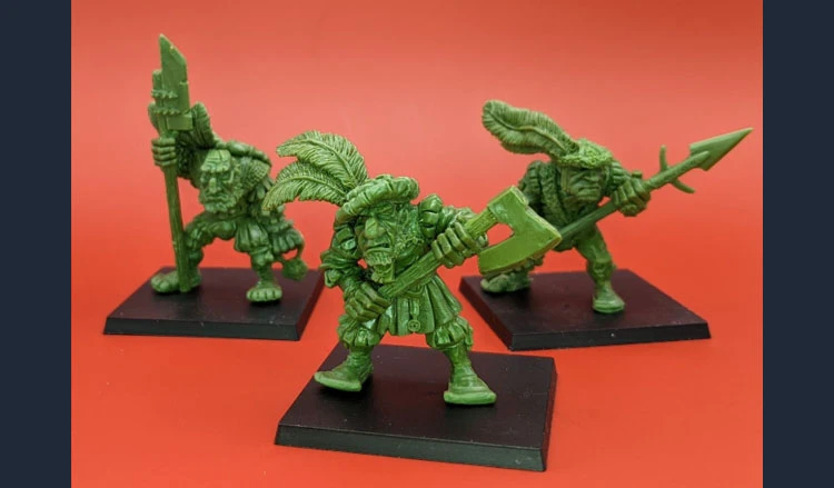 Three green ogre miniatures are displayed against a bright red background. Each figure is uniquely posed: one holds a large axe, another wields a spear, and the third carries a staff adorned with feathers. They stand on black bases, showcasing their detailed sculpting that pays homage to classic oldhammer aesthetics. The miniatures exhibit exaggerated features and playful expressions, embodying a whimsical yet menacing presence suitable for wargames and RPGs.