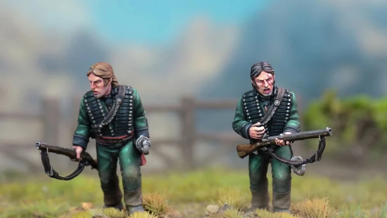 Two detailed miniatures of soldiers from the Sharpe’s Rifles series are displayed on textured bases, set against a blurred natural background. Both figures wear green uniforms with black crossbelts; one holds a rifle and has a serious expression, while the other appears to be in a more contemplative pose. The bases feature tufts of grass, enhancing the realistic terrain effect.