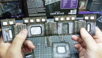 A hand holds a piece of unpainted cardboard terrain with a rectangular window and weathered metal texture. On a green cutting mat, additional assembled cardboard terrain pieces with similar windows, ladders, and industrial details are visible. In the background, a product box for "CORE SPACE" by Battle Systems is partially visible, with text and a barcode.