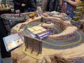 A detailed tabletop gaming setup features a winding race track made of textured terrain, resembling rocky cliffs and sandy surfaces. Miniature vehicles are positioned along the track, with various game components, including dice and rulebooks, scattered on the table. In the background, several people are engaged in the game, adding to the lively atmosphere of the event.