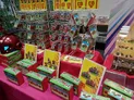 A vibrant display of miniature wargaming products is showcased on a table covered with a bright pink cloth. Various boxes of miniatures are arranged neatly, with some priced at £9 and others at £15, featuring colorful artwork on their packaging. Behind the boxes, additional miniatures are displayed in clear packaging, highlighting a diverse selection for wargaming enthusiasts.