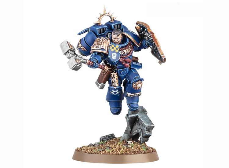 A dynamic miniature of a Space Marine captain in blue armor, poised mid-action with a thunder hammer in one hand and a relic shield in the other. He features a jump pack on his back and has a determined expression, with details like a skull emblem on his chest and a decorative shoulder pad. The base is textured with dirt and small rocks, adding to the overall dramatic presentation.