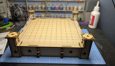 A wooden Rumble Slam Rookie Ring is displayed, featuring a square mat with a grid pattern and four corner posts adorned with star designs. The ring is surrounded by white ropes, and the base has a simple, clean design with additional star motifs. In the background, various paint pots are visible, indicating a workspace for miniature painting and modeling.