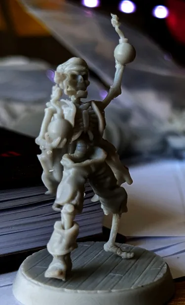 A detailed miniature of a skeleton pirate stands with one arm raised, holding a round object, possibly a cannonball or a drink. The figure is intricately sculpted, showcasing skeletal features and pirate attire, including tattered clothing. The background is slightly blurred, revealing stacks of game-related materials, suggesting a tabletop gaming environment.