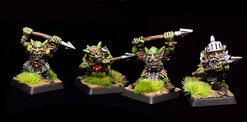 Four dynamic goblin miniatures are displayed, each wielding a spear. They are painted in vibrant greens with various armor details, including chainmail and leather, showcasing their distinct personalities. The goblins stand on textured bases adorned with tufts of grass, enhancing their lively appearance. One goblin wears a helmet, while another has a red tunic, adding variety to their designs. The overall scene captures the whimsical and mischievous essence typical of Kev Adams' goblin creations.