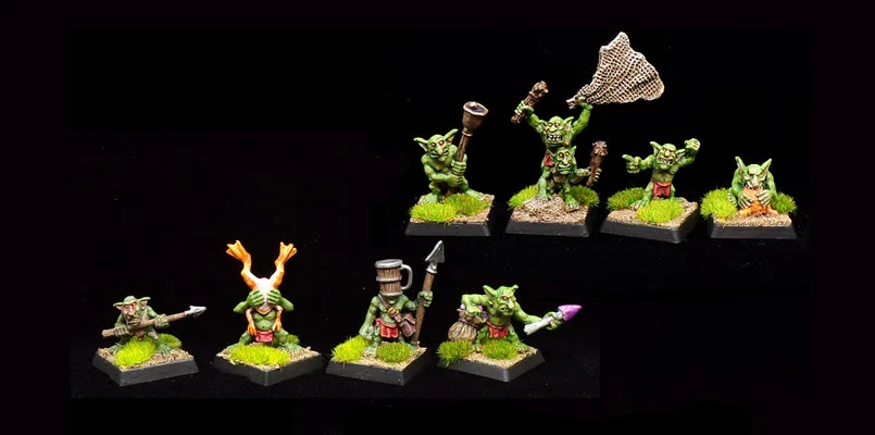 The image features a collection of painted goblin miniatures, showcasing their vibrant green skin and dynamic poses. There are eight goblins displayed on textured bases with tufts of grass, each holding various weapons and items, such as a spear, a net, and a mug. The goblins exhibit a range of expressions and actions, emphasizing their character and personality, typical of Kev Adams' sculpting style.