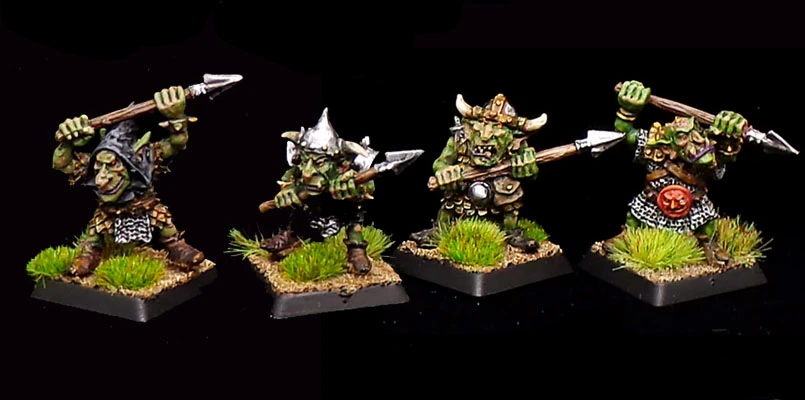 Four dynamic goblin miniatures are displayed, each wielding a spear and standing on textured bases adorned with tufts of grass. The goblins exhibit a variety of armor styles, including a hooded figure, one with a horned helmet, and another with a shield featuring a red emblem. Their skin is a vibrant green, and they have expressive facial features that convey a sense of mischief and character. The overall scene captures the whimsical and detailed artistry typical of Kev Adams' goblin designs.